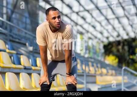 Müde hispanische Athleten, die sich nach dem Joggen gebeugt und atmet, Mann, der an sonnigen Tagen Sport treibt, mit aktiven Übungen und Fitness, Sportler, der sich nach dem Joggen ausruht. Stockfoto