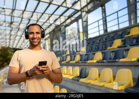 Sportler mit Telefon lächelnd und mit Blick auf die Kamera, Mann am Morgen joggen und trainieren im Sportstadion, Mann hört Musik in Kopfhörern und Podcasts Online-Radio. Stockfoto