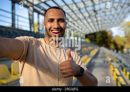 Nahaufnahme. . Junger afroamerikanischer Sportler steht im Stadion mit Kopfhörern, macht ein Selfie am Telefon, spricht bei einem Videoanruf. Zeigt der Kamera eine Supergeste an. Stockfoto