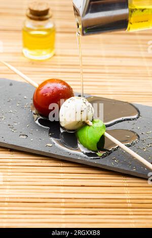 Extra natives Olivenöl über einen Salatspieß gießen. Tomaten, Mozzarella-Käse und Basilikumblätter auf einem Tablett. Stockfoto