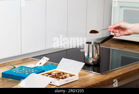 Zürich, Schweiz - Januar 7 2023: Lindt Mini Pralines Box. Offene kleine Geschenkbox mit Schweizer Schokoladensorten und eine Hand junger Frau, die türkischen Kaffee auf der Küchenplatte macht. Stockfoto