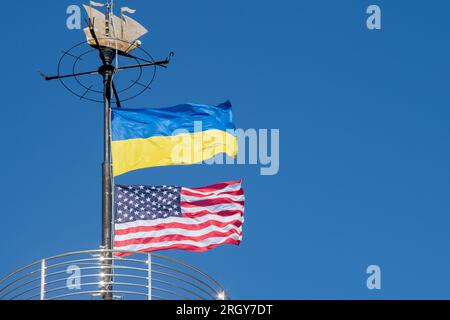 Die Flaggen der Ukraine und der USA flattern auf einem Fahnenmast vor einem blauen Himmel. Statussymbol. Ukrainische blaue und gelbe Flagge. Windtag im Freien. Stockfoto