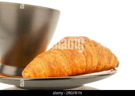 Ein frisch gebackenes, duftendes Croissant auf einer Untertasse mit schwarzem Becher, Nahaufnahme, isoliert auf Weiß. Stockfoto