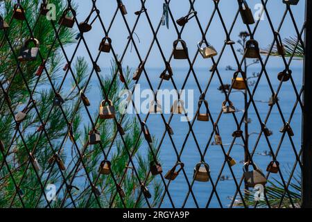 Ein Maschenzaun am Meer mit Vorhängeschlössern, die die Liebe symbolisieren. Liebesschlösser werden in großer Zahl an der Meerespromenade am Zaun aufgehängt. Meer im Hintergrund Stockfoto