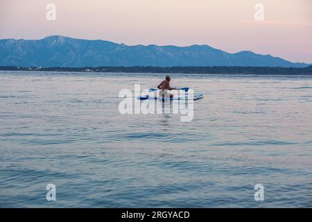 Ein gealterter Bär und grauer Mann schwimmt bei Sonnenuntergang auf einem sack im Meer. Schöne ruhige Reinigung des Meeres und der Berge im Hintergrund. Stockfoto
