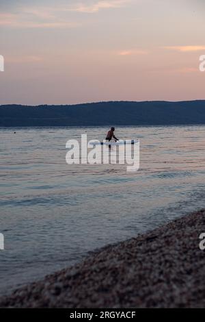 Ein gealterter Bär und grauer Mann schwimmt bei Sonnenuntergang auf einem sack im Meer. Schöne ruhige Reinigung des Meeres und der Berge im Hintergrund. Stockfoto