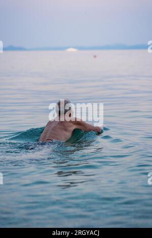 Der alte Mann schwimmt in der Adria. Ein Mann mit grauen Haaren schwimmt im ruhigen blauen Wasser des Meeres bei Sonnenuntergang. Stockfoto