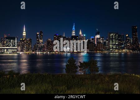 Die ikonische Skyline von Manhattan von Long Island City aus gesehen. Bäume im Vordergrund, Lichter der Stadt in der wolkenlosen Sommernacht. Stockfoto