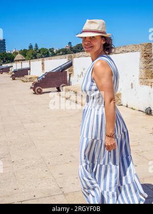 Lächelnder kaukasischer Tourist, der ein Sommerkleid und einen alten Strohhut trägt und auf einer antiken Latinamerikanischen Yachthafenpromenade spaziert. Stockfoto