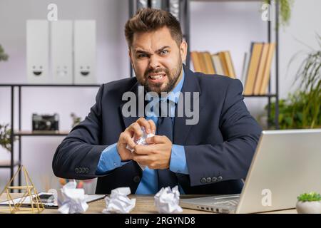 Wütender, wütender junger Mann, der im Büro arbeitet, zerknittert Papier wirft, Nervenzusammenbruch bei der Arbeit hat, Migräne, Kopfschmerzen, Stressbewältigung, psychische Probleme, Sie verlieren die Beherrschung, reagieren auf Fehler Stockfoto