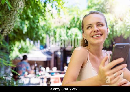 Glückliche junge Frau, die im Sommer im Freiluftcafé sitzt und ihr Handy in der Hand hält. Stockfoto