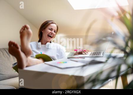 Eine junge Studentin sitzt zu Hause am Schreibtisch mit den Füßen auf dem Tisch und amüsiert sich, lacht, während sie ihren Laptop benutzt. Stockfoto