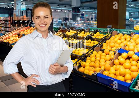 Eine junge Frau, die Filialleiterin im Verkaufsbereich vor Kisten mit Obst, Zitronen und Orangen steht. Stockfoto