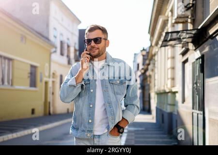 Ein junger Mann in Jeanskleidung und Sonnenbrille, der die Straße entlang läuft und telefoniert Stockfoto