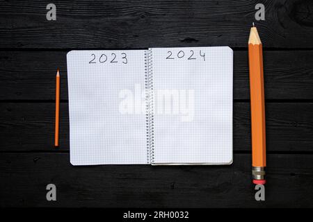 Das Jahr 2023 und 2024 steht auf den Seiten eines Notizbuchs und daneben liegt ein großer und kleiner Bleistift auf einem schwarzen Tisch, das Ende des alten Jahres an Stockfoto