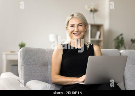 Glückliche, wunderschöne blonde, reife, freiberufliche Geschäftsfrau, die auf der Couch sitzt Stockfoto