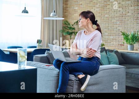 Eine ernsthafte Frau mittleren Alters, die zu Hause auf dem Sofa auf dem Laptop sitzt Stockfoto