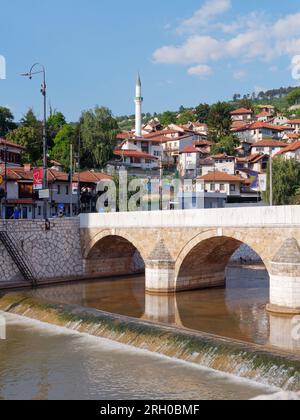 Brücke über den Fluss Miljacka mit Häusern und Moschee dahinter in Sarajevo, Bosnien und Herzegowina, 12. August 2023. Stockfoto