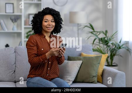 Porträt einer jungen erfolgreichen Frau zu Hause, einer afroamerikanischen Frau, die lächelt und in die Kamera schaut, das Telefon in der Hand hält, während sie auf der Couch im Wohnzimmer sitzt. Stockfoto