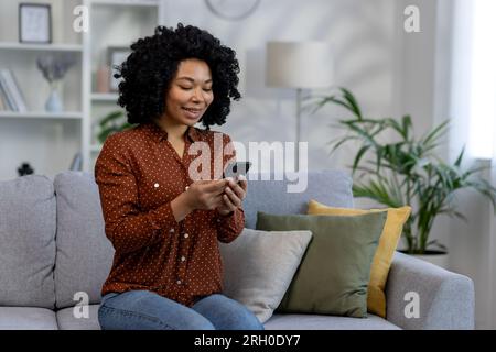 Junge, wunderschöne afroamerikanische Frau, die zu Hause auf dem Sofa sitzt, Frau mit lockigem Haar, die das Telefon in den Händen hält, die Online-Anwendung im Wohnzimmer nutzt, Internetseiten durchsucht und Nachrichten tippt Stockfoto