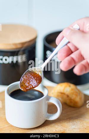 Braune Zuckerkörner ziehen den heißen Kaffee zu einer sirupen Konsistenz auf, bevor sie in einen frisch gebrühten schwarzen Espresso gerührt werden, der trinkfertig und heiß ist Stockfoto