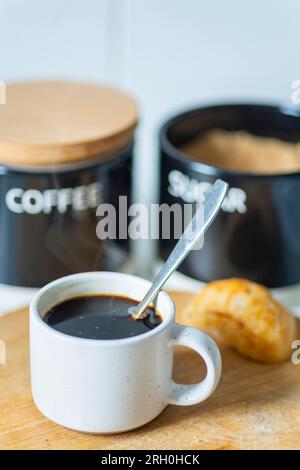 Frisch gegossen, heiß, trinkfertig, in einem Espresso-Kaffeebecher und Silberlöffel zum Rühren, auf einem Holzbrett neben einem Brötchen, Zucker und CO Stockfoto