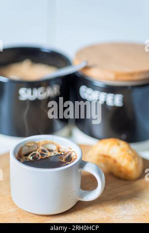 Frisch gegossen und dampfend heiß, trinkfertig, Kaffeebecher auf einem Holzbrett neben einem Brötchen, Demerarazucker und Kaffeebehälter, kürzlich Stockfoto