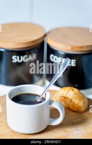 Frisch gegossen, heiß, trinkfertig, in einem Espresso-Kaffeebecher und Silberlöffel zum Rühren, auf einem Holzbrett neben einem Brötchen, Zucker und CO Stockfoto