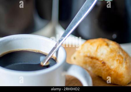 Frisch gegossen, heiß, trinkfertig, in einem Espresso-Kaffeebecher und Silberlöffel zum Rühren, auf einem Holzbrett neben einem Brötchen, Zucker und CO Stockfoto