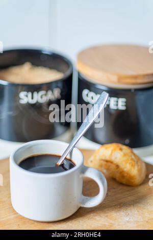 Frisch gegossen, heiß, trinkfertig, in einem Espresso-Kaffeebecher und Silberlöffel zum Rühren, auf einem Holzbrett neben einem Brötchen, Zucker und CO Stockfoto