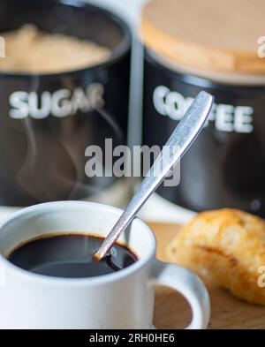 Frisch gegossen, heiß, trinkfertig, in einem Espresso-Kaffeebecher und Silberlöffel zum Rühren, auf einem Holzbrett neben einem Brötchen, Zucker und CO Stockfoto