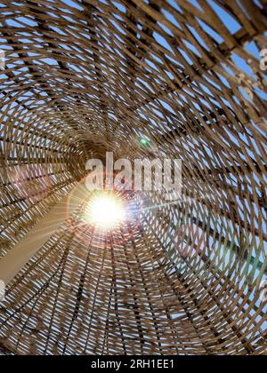 Strohstrukturierter Rattanschirm am Strand, vertikal. Sonnenstrahl fällt an sonnigen Tagen durch einen exotischen Sonnenschirm. Sommerspielhintergrund mit Kopierraum Stockfoto