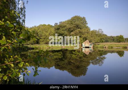 Aufgenommen an einem sonnigen Sommertag mit klarem blauen Himmel, zeigt es einen alten Bootshaus auf einem Pool, der sich im Wasser spiegelt Stockfoto