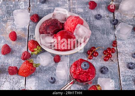 Kugeln aus selbstgemachter Erdbeere, roter Johannisbeere, Himbeere, Heidelbeereis, Sorbet in Schale, Kugeln, Beeren und Eiswürfel über auf blauem Holzbackgroun Stockfoto