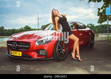 Roter Mercedes AMG-GTS Sportscar (2015), der auf einer Straße mit einer stilvollen Frau geparkt ist, ein kurzes schwarzes Kleid trägt und hohe Absätze trägt und sich gegen das Auto lehnt Stockfoto