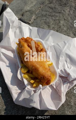 Fish and Chips, zerstoßener Kabeljau, in einem Tablett, das mit einer Holzgabel am Strand in Papier eingewickelt ist. Traditionelles britisches Speisenkonzept Stockfoto
