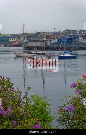 Ein hölzernes Langboot und Yachten, die an einem bedeckten frühen Morgen im Juni im Hafen von Stornoway auf der Isle of Lewis in den Hebriden festgemacht wurden. Stockfoto