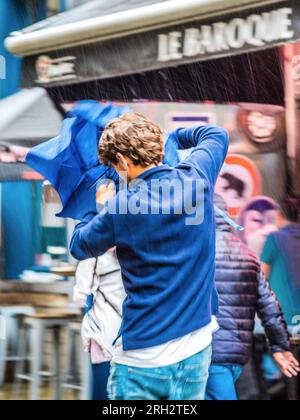 Junger Mann, der Schwierigkeiten hat, bei Regen einen kaputten Regenschirm zu öffnen (langsame Verschlusszeit/Bewegungsunschärfe) – Brüssel, Belgien. Stockfoto