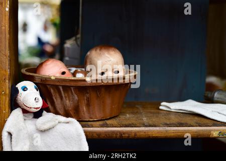 Auf einem Flohmarkt schauen die Puppenköpfe mit leeren Augenhöhlen über dem Rand einer Backform, flankiert von einem Puppenhund Stockfoto