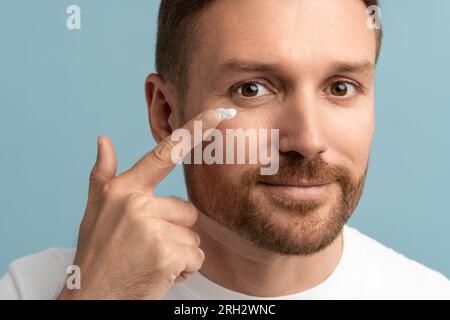 Nahaufnahme eines bärtigen gutaussehenden Mannes, der feuchtigkeitsspendende Creme auf die Gesichtshaut aufträgt und in die Kamera schaut. Wellness Stockfoto