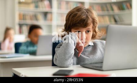 Fokussierter weißer Schuljunge, der am Schreibtisch sitzt und in einem Grundschulraum einen Laptop benutzt Stockfoto