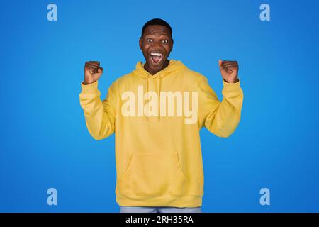 Fröhlich überraschter junger afroamerikanischer Mann im Sweatshirt erhebt die Hände und feiert den Sieg Stockfoto