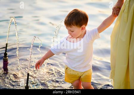 Ein kleiner Junge und seine mutter ruhen sich aus, indem sie an einem heißen Sommertag im kühlen Wasser des Brunnens spazieren gehen. Kind im Alter von etwa zwei Jahren (ein Jahr elf mont Stockfoto