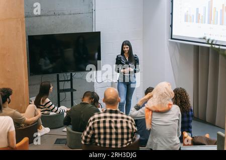 Junge und schöne Geschäftsfrau lächelt und präsentiert einer vielfältigen Gruppe von Menschen in einem kleinen Vorlesungssaal ein Diagramm Stockfoto