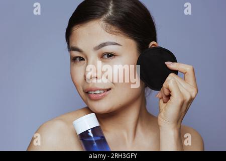 Moderne asiatische Frau mit Make-up-Entferner und schwarzem Baumwollkissen auf blauem Hintergrund. Stockfoto