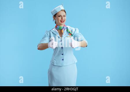 Lächelnde, stilvolle Stewardess auf blauem Hintergrund in blauer Uniform mit Daumen nach oben. Stockfoto