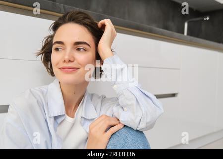 Das Porträt einer jungen Frau sitzt auf dem Boden, lächelt und schaut aus dem Fenster, genießt die Freizeit zu Hause, entspannt sich, hat einen traumhaften Gesichtsausdruck Stockfoto