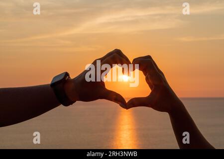 Hände Herz Meer sanset. Hände, die eine Herzform bilden, die vor dem Sonnenhimmel eines Sonnenaufgangs oder Sonnenuntergangs am Strand liegt Stockfoto