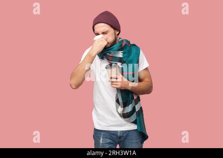 Porträt eines ungesunden bärtigen Mannes in weißem T-Shirt, Bohnenhut und Schal stehen, Pappbecher halten und niesen, sich erkälten, laufende Nase. Studio-Aufnahme im Innenbereich isoliert auf pinkfarbenem Hintergrund. Stockfoto