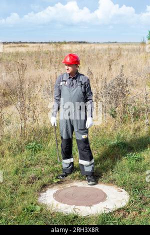 Ein Arbeiter in Overalls und einem Helm in der Nähe eines Mannlochs. Kontrolle und Instandhaltung von Kanalisation, Klärgruben und Rohrleitungen. Stockfoto